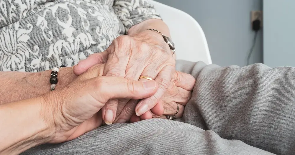Mãos de uma pessoa segurando as mãos de um idoso no colo, cena de cuidado ao escolher um cuidador para um idoso
